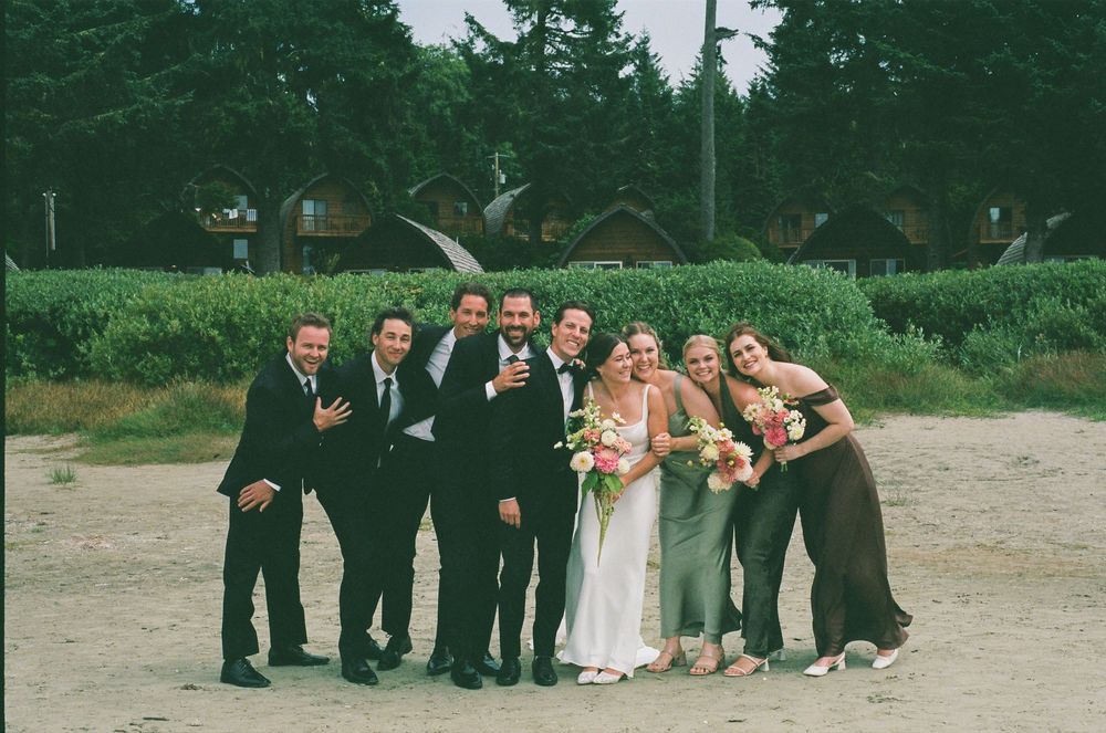 Wedding party posing outdoors, bride in white gown with bridesmaids and groomsmen in formal attire, green hedges background.