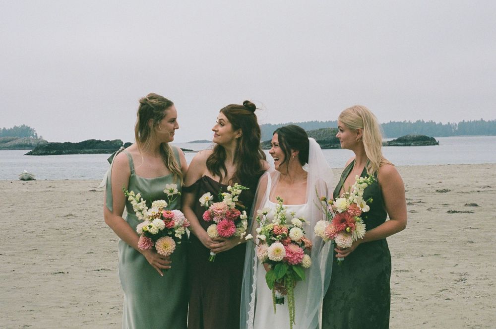 Four bridesmaids in sage green dresses holding floral bouquets on a beach at a coastal wedding ceremony.