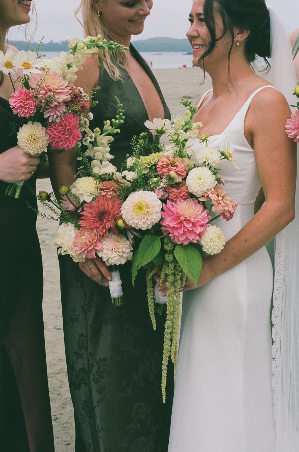 Bride holding lush pink dahlia and white wedding bouquet with bridesmaids in dark green gowns on beach.