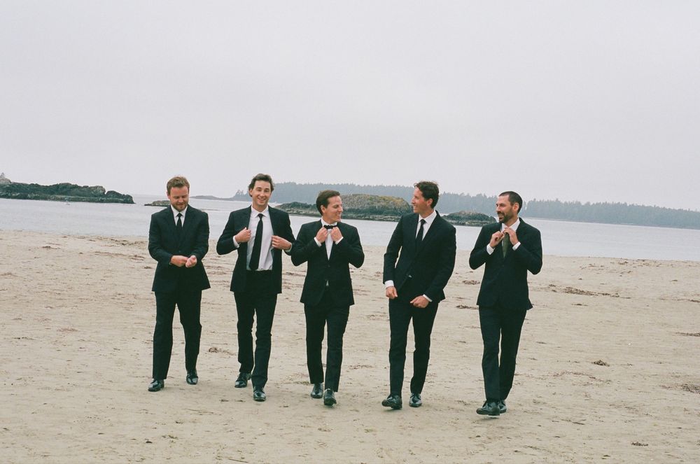 Five groomsmen in black suits walking on a sandy beach, adjusting ties and laughing together.