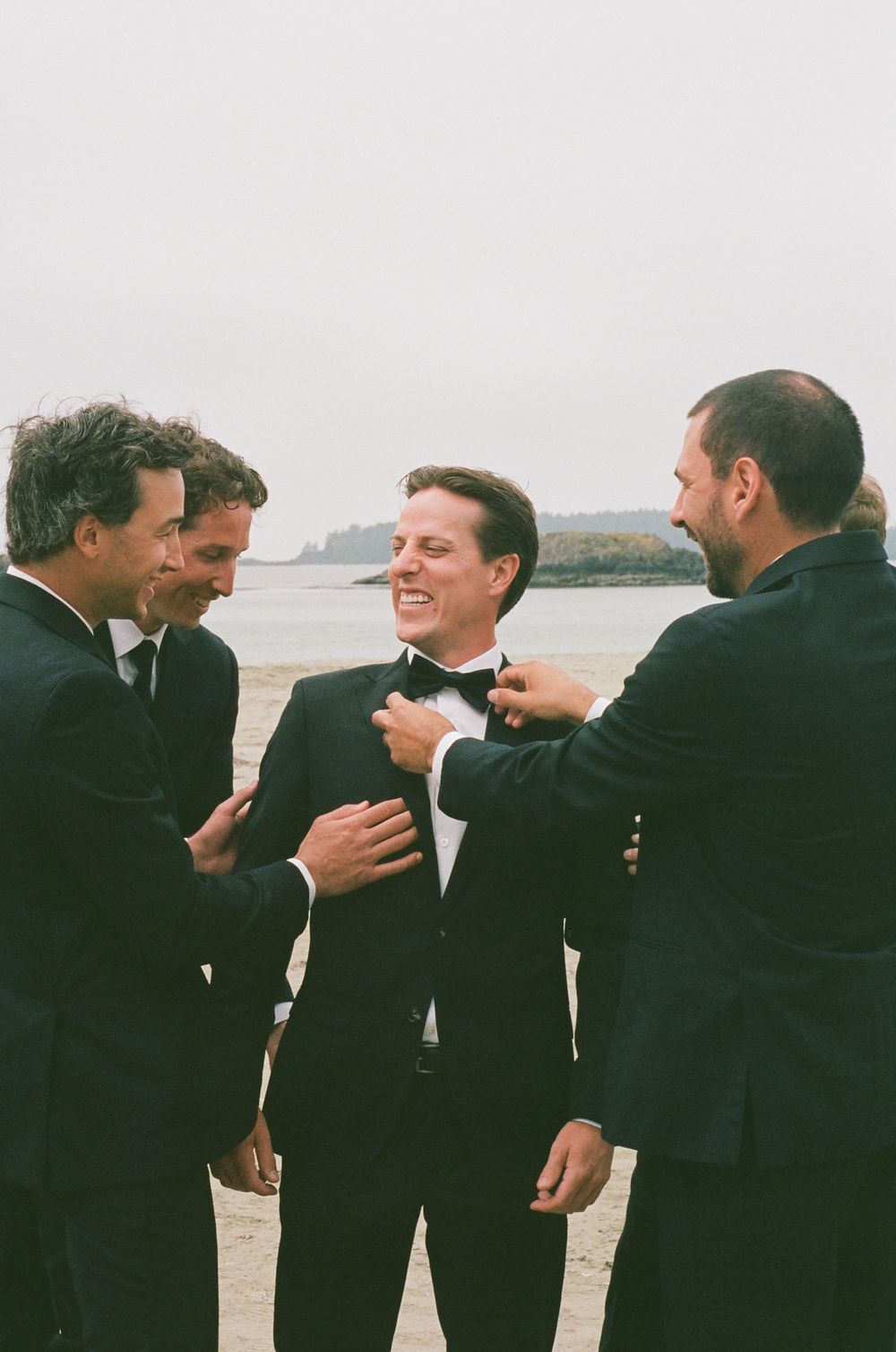 Groomsmen in black tuxedos laughing and adjusting the grooms bow tie on a beach wedding day.