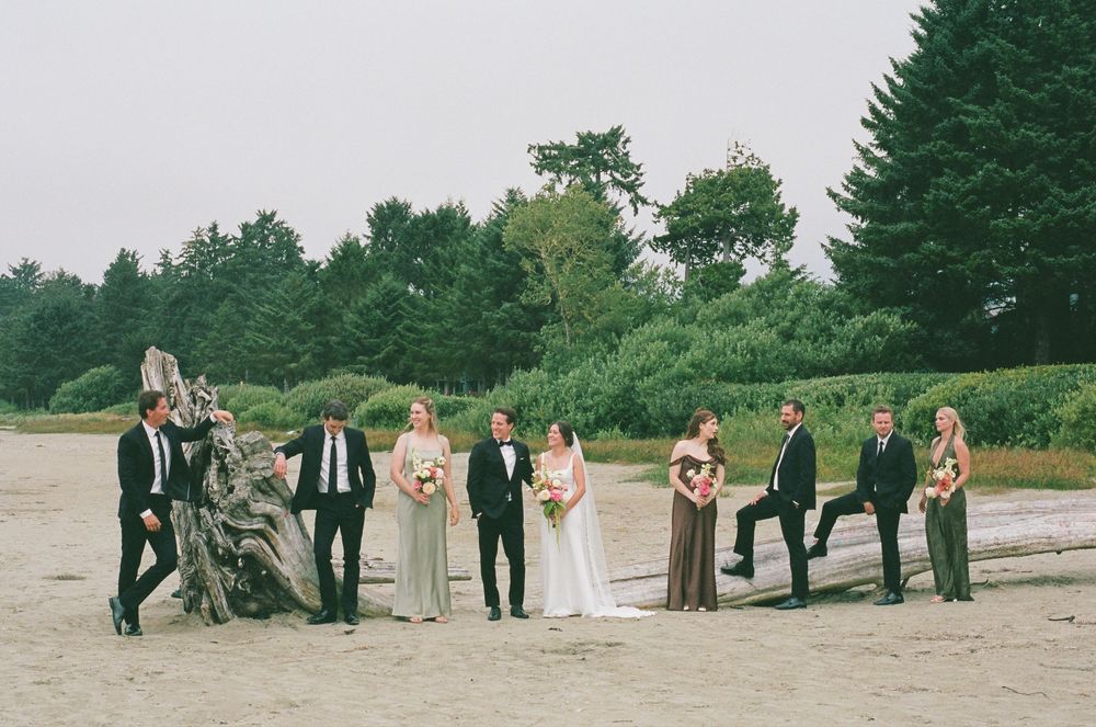 Wedding party posing on sandy beach with driftwood and evergreen trees backdrop.