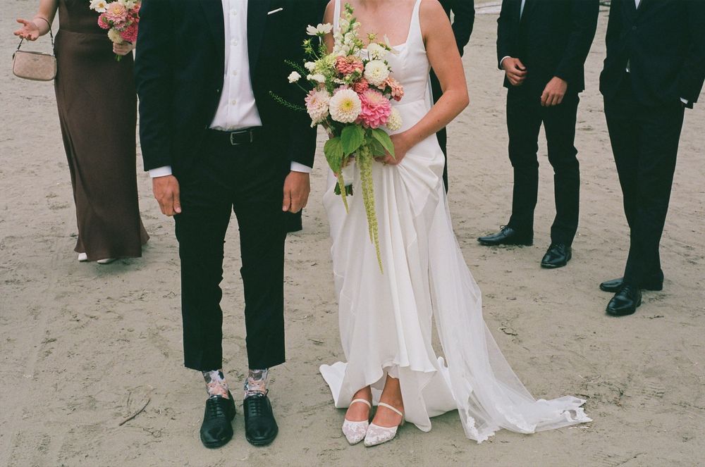 Beach wedding couple, bride holding pink and white bouquet, groom in black suit with fun socks, bridal party behind them.