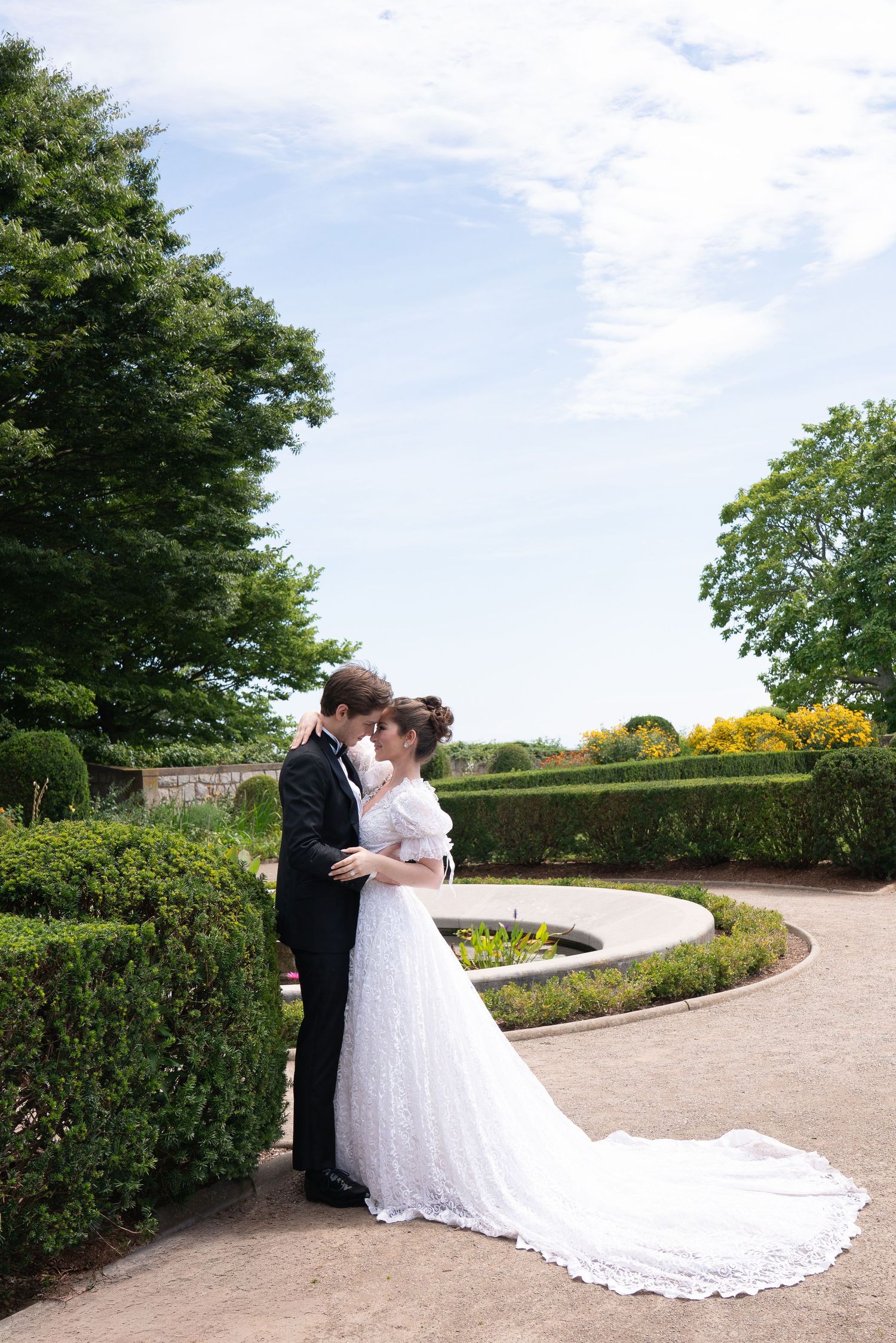 Bride and groom share a romantic kiss in a formal garden with manicured hedges and blue sky backdrop.