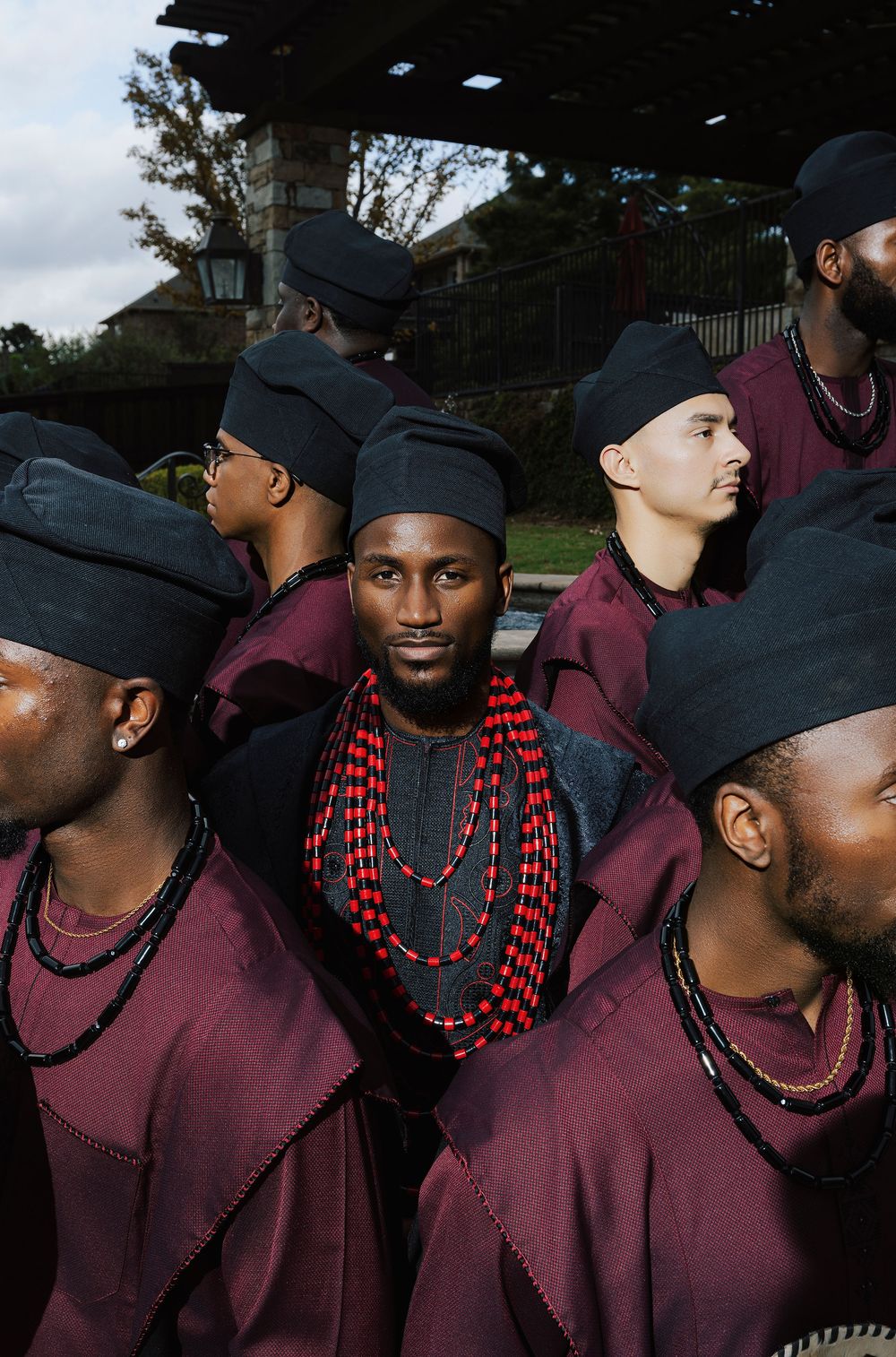 Group of men in burgundy robes and black caps, with one man wearing layered red beaded necklaces, standing outdoors.