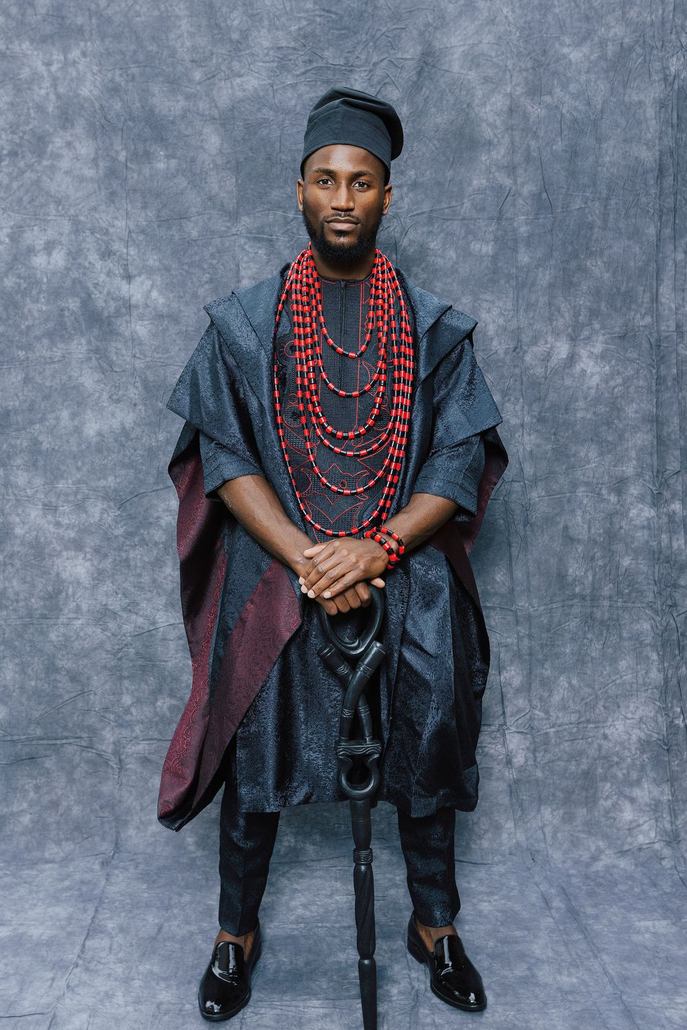 Man in traditional African attire with red beaded necklaces and dark agbada robe posing for a portrait.