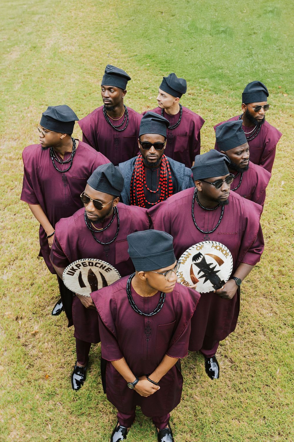 Group of men in matching burgundy traditional African attire and black caps posing outdoors on green grass.
