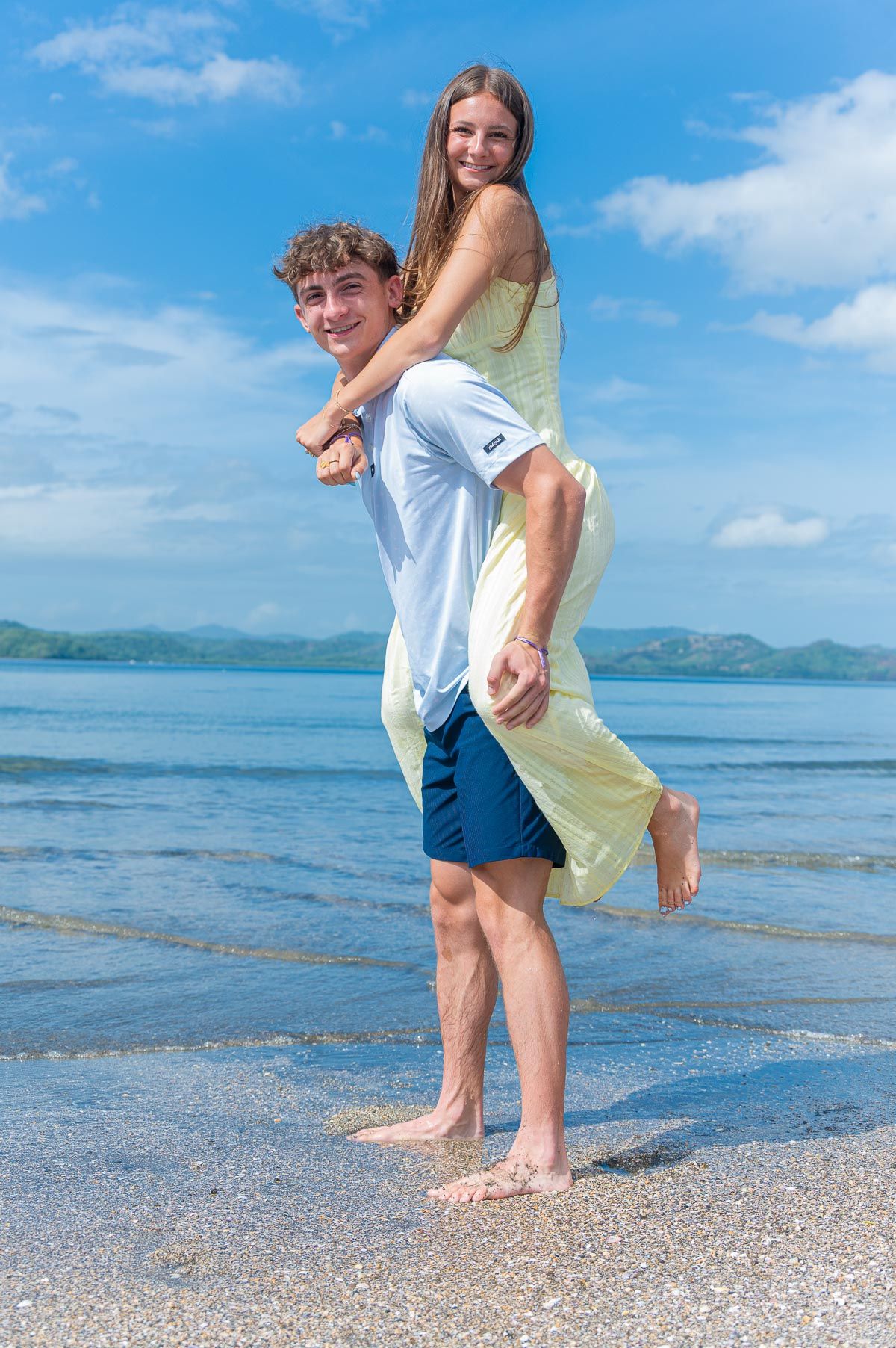 Young couple enjoying a piggyback ride on a tropical beach, with clear blue skies and ocean waves in the background.