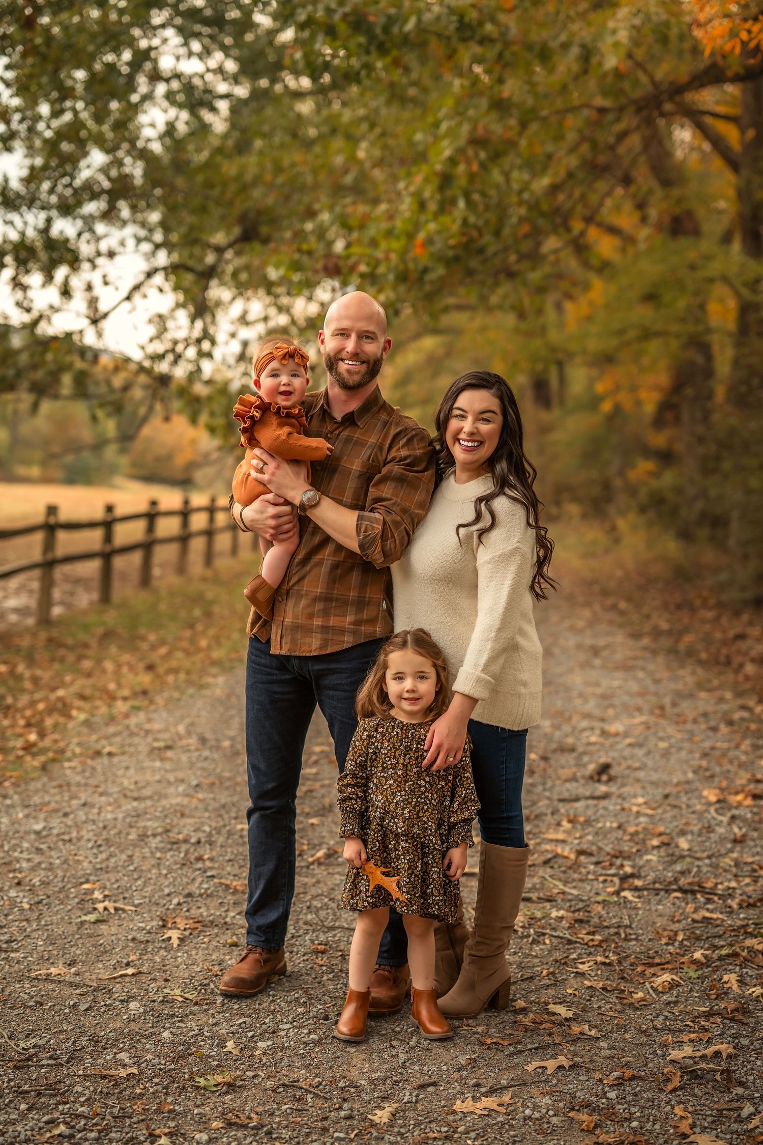 Family of four posing on a scenic autumn path surrounded by colorful fall foliage and a wooden fence.