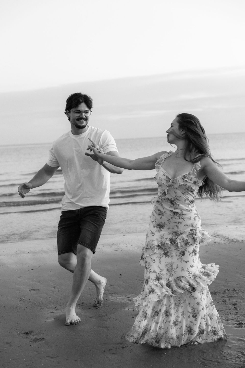 Black and white photo of a happy couple running barefoot on a beach, woman in floral dress, man in white tee and shorts.