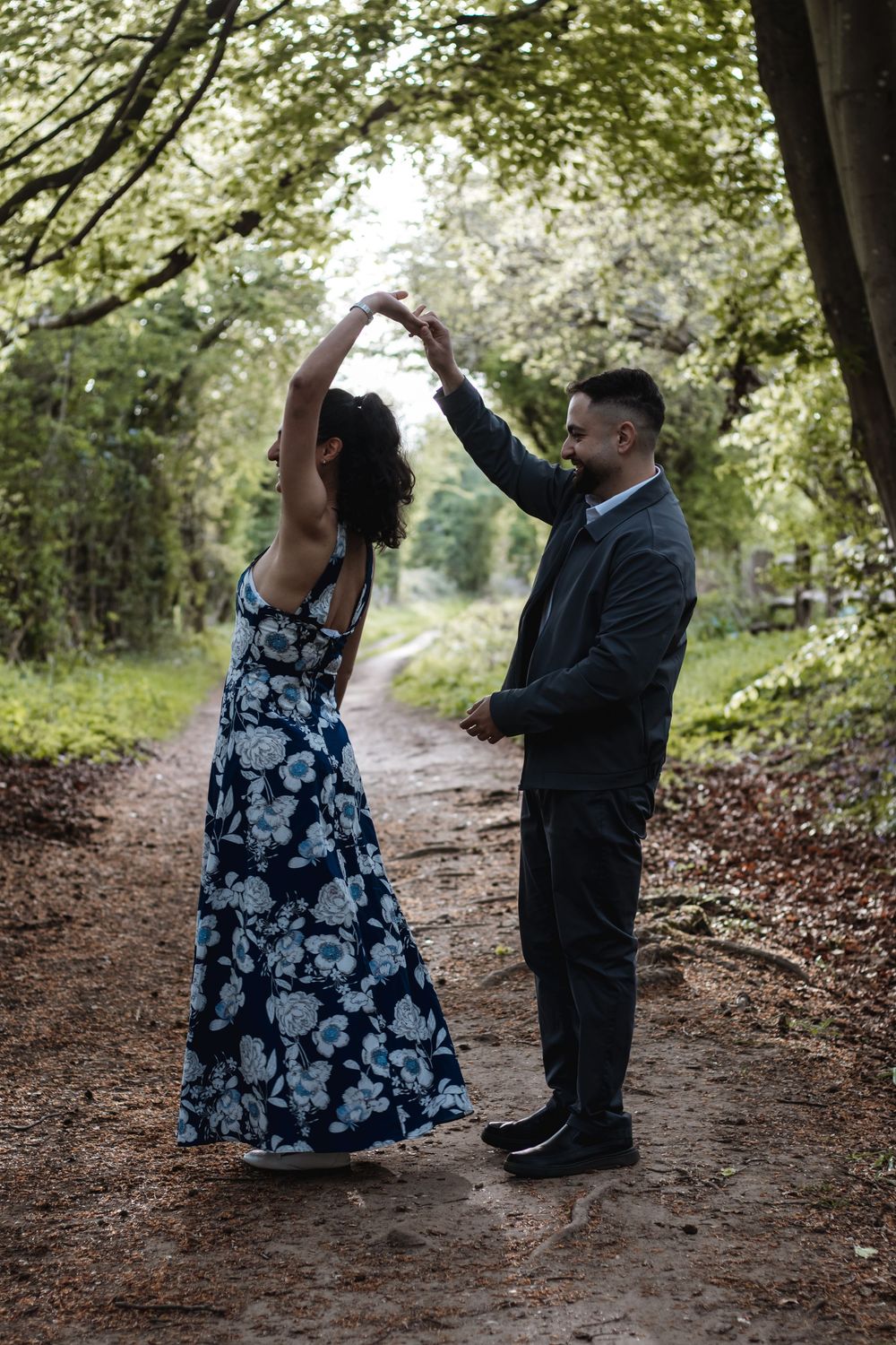 Couple dancing on a forest path, woman in floral dress being twirled by man in dark suit.
