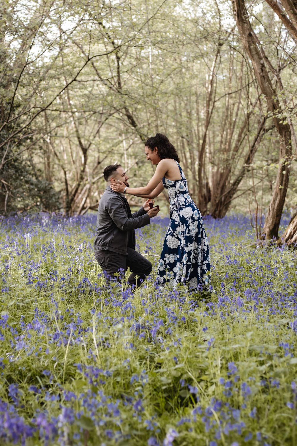 Man proposes to woman in a bluebell forest, kneeling among purple wildflowers in a romantic outdoor setting.