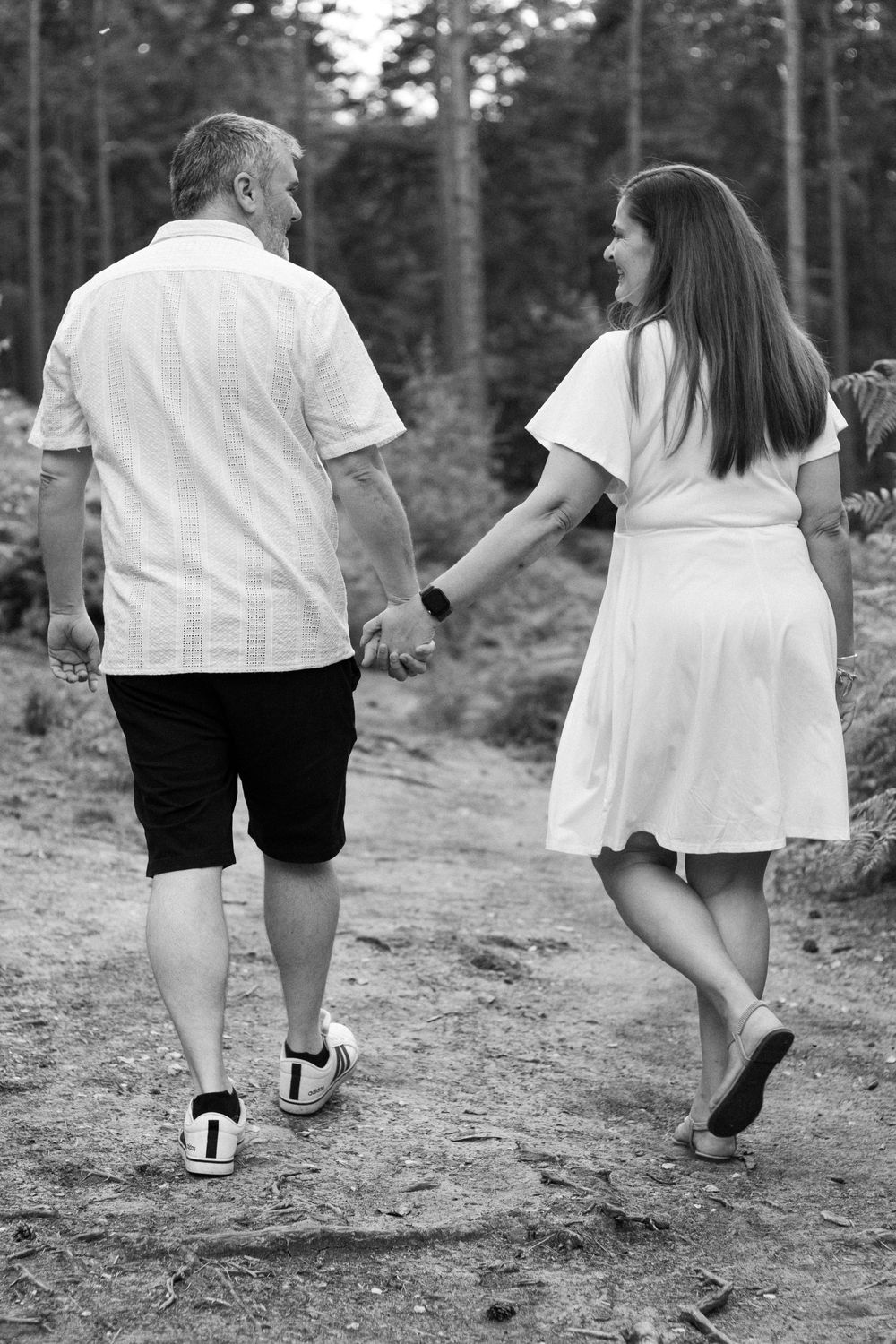 Black and white photo of a couple holding hands while walking on a forest path, woman in white dress looks back smiling.