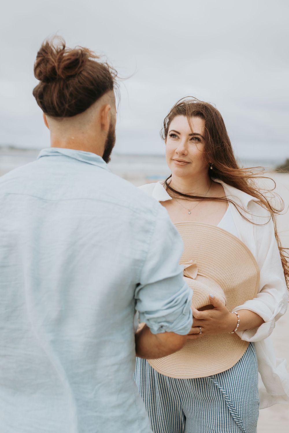 Couple on a windy beach, woman in white shirt holds straw hat, man in blue linen shirt faces her.