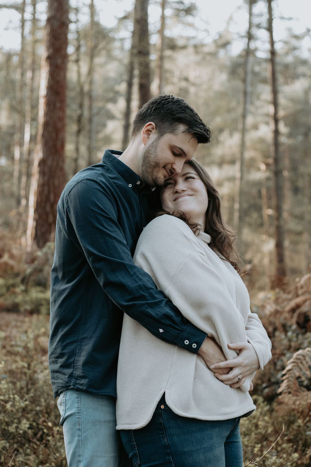 A couple embraces tenderly in a forest, the man holding the woman from behind as she smiles contentedly.