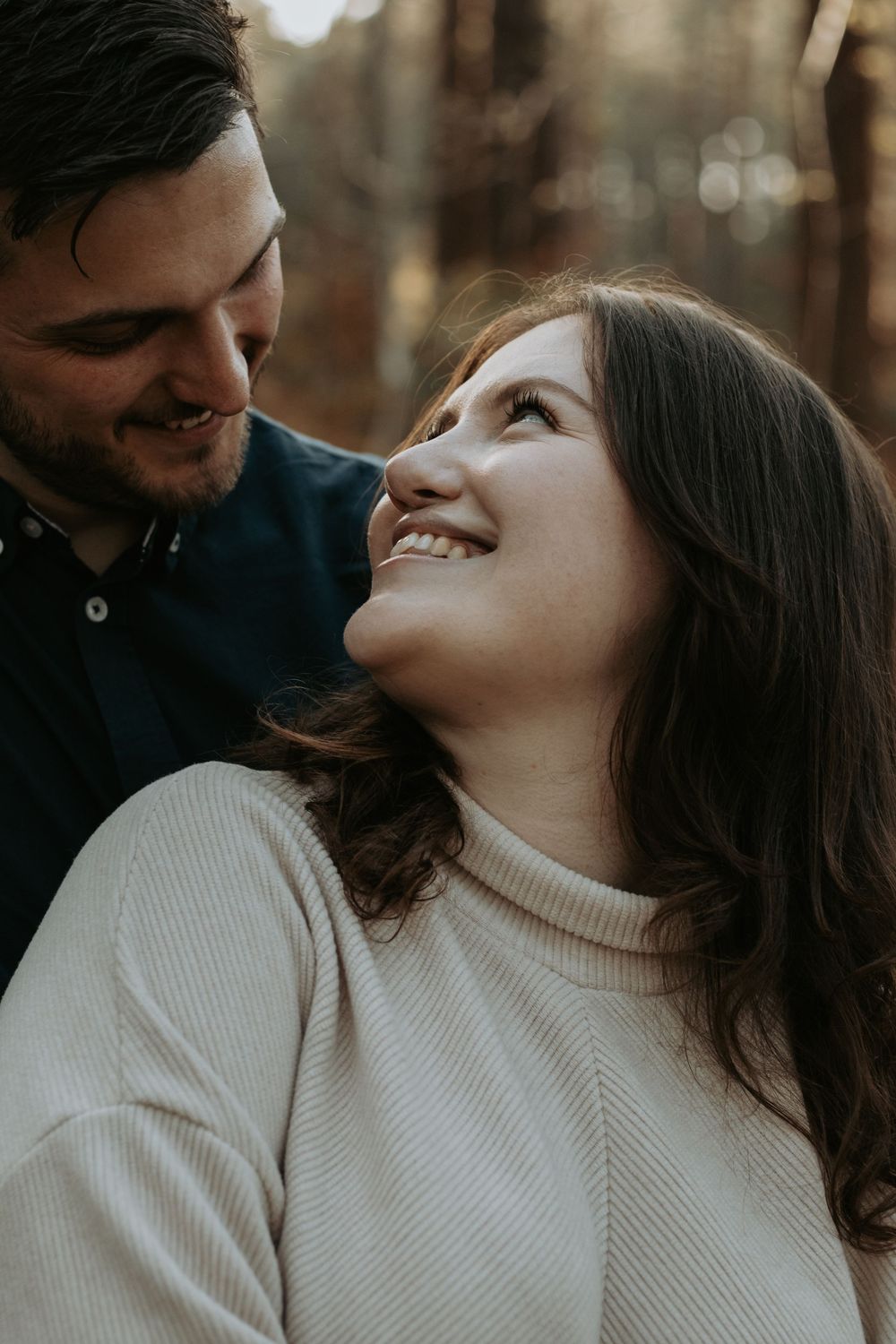 A smiling woman in a cream sweater gazes up at a man in a blue shirt, sharing a romantic moment outdoors.