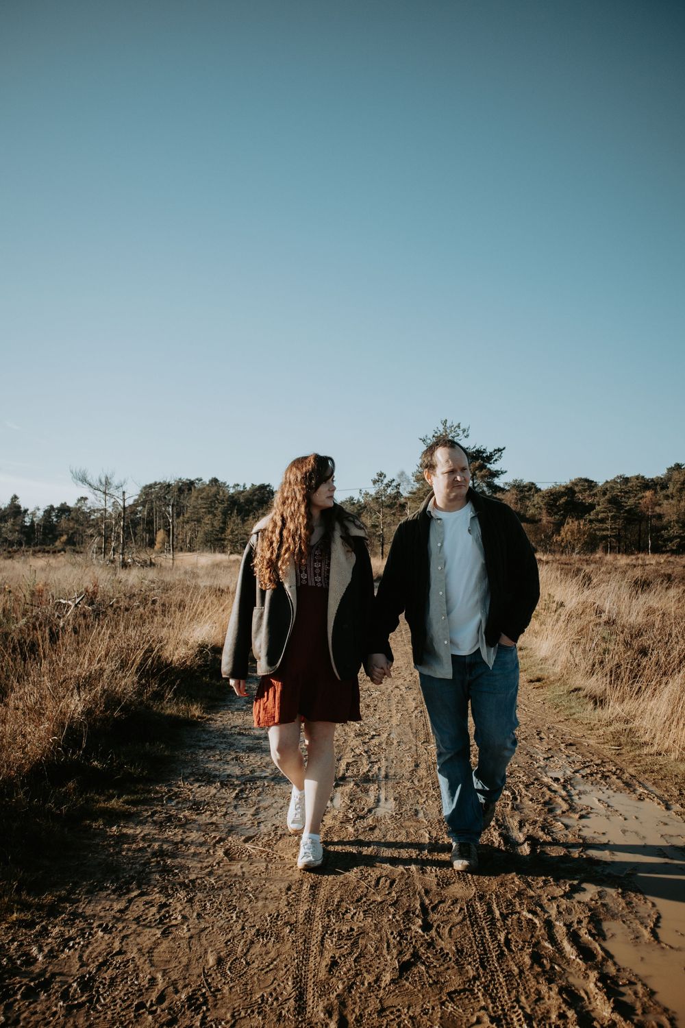 Couple holding hands walking on a muddy dirt path through golden fields under a clear blue sky.