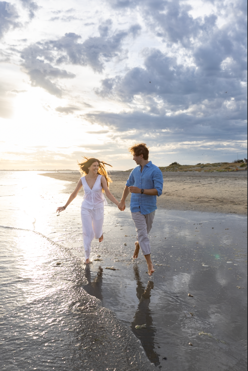 Couple holding hands running barefoot on a sunny beach at sunset with dramatic cloudy sky.