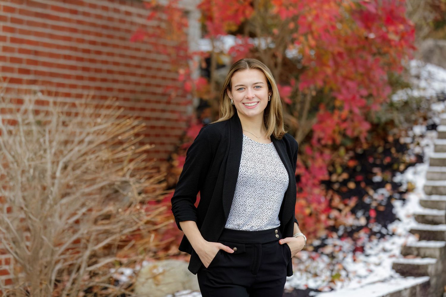 Young woman smiling in black blazer and gray top, posing outdoors near brick wall with red autumn foliage.