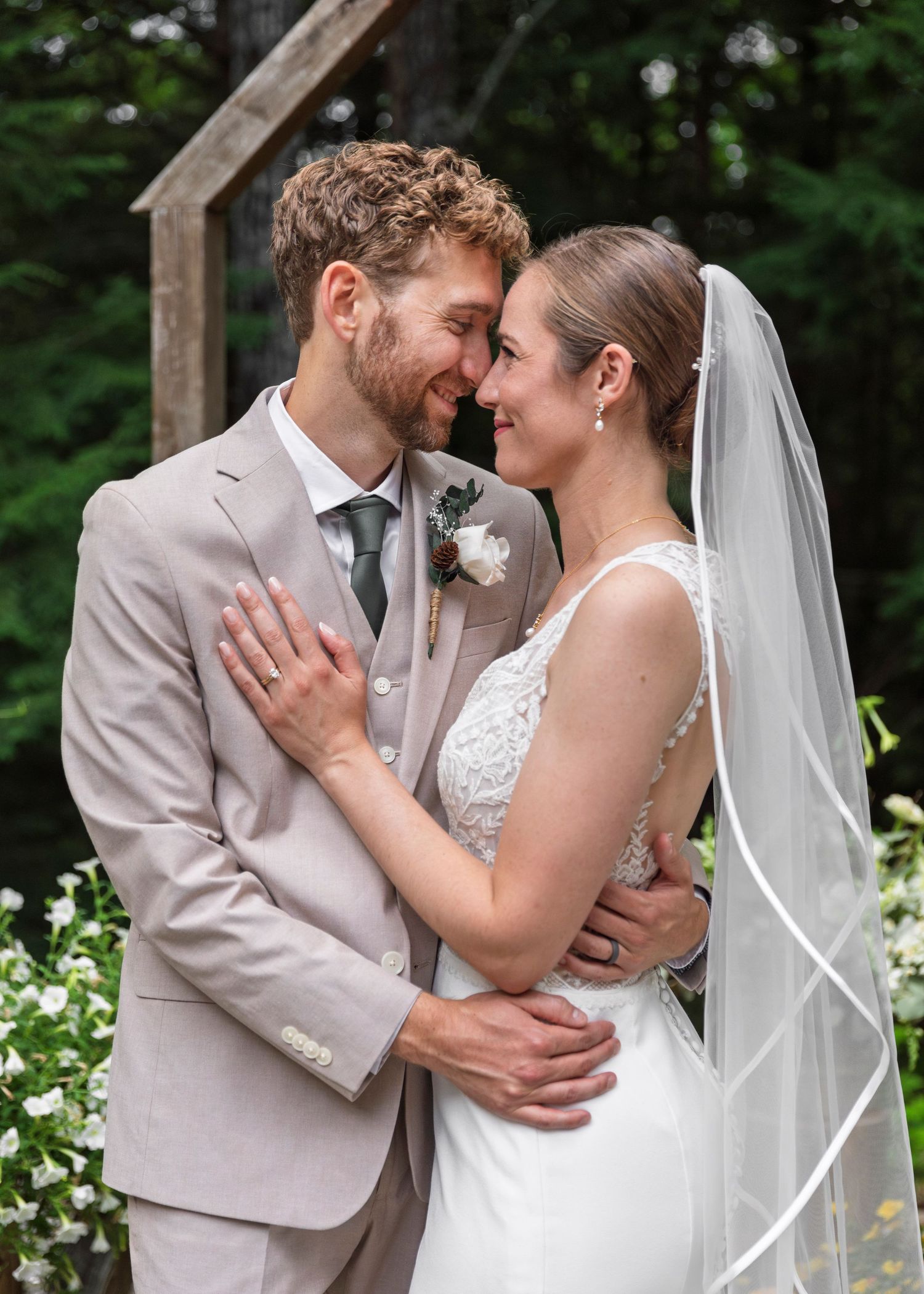 A bride and groom share a romantic forehead touch outdoors, she in a lace veil, he in a tan suit.