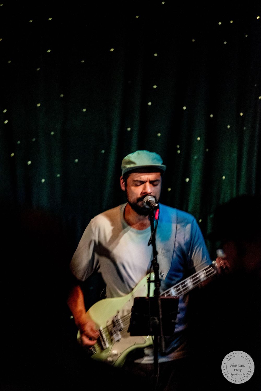 Man in cap playing white electric guitar and singing into microphone on stage with green curtain backdrop.