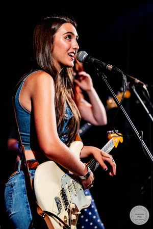 Female guitarist singing into microphone on stage holding a white Fender guitar at a live concert.