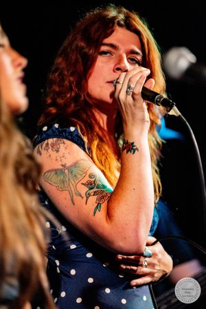 Tattooed female singer with red curly hair performing live on stage holding microphone in blue polka dot dress.
