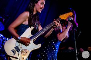 Young woman playing white electric guitar on stage, smiling, with another performer singing into microphone behind her.
