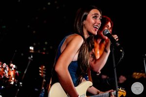 Female guitarist singing passionately on stage, holding white electric guitar under concert lights.
