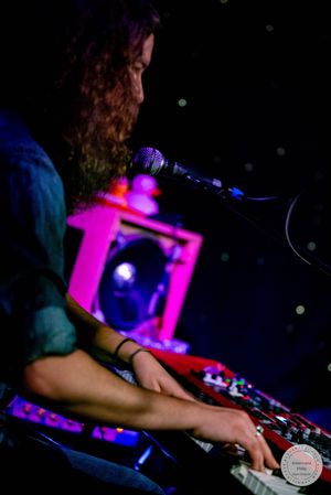 Curly-haired musician plays synthesizer keyboard on stage near microphone under pink and purple concert lighting.