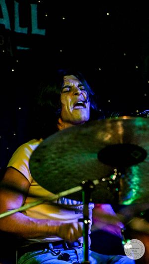 Drummer performing live on stage under colorful lights, playing cymbals with drumsticks in a dark concert venue.