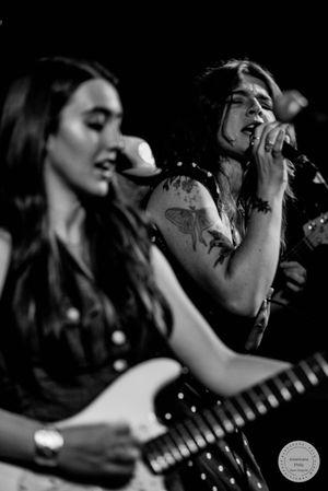Two female musicians perform on stage in black and white photo, one singing into mic with tattoos visible.