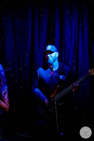 Male bassist in cap smiles on stage under blue concert lighting in front of dark curtain backdrop.