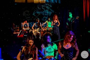 Band performing on a colorful lit stage at a music hall, with female singers in front and guitarists in the background.