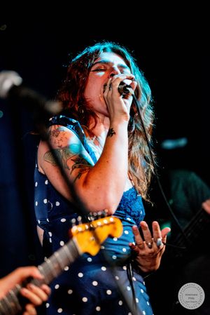Female singer in blue polka dot dress performing live on stage while holding microphone with tattoos visible.