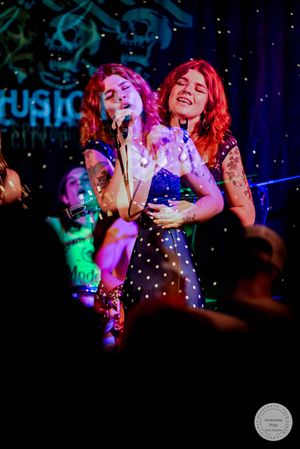 Two tattooed female singers sharing microphone on stage in matching blue polka dot dresses at live music venue.