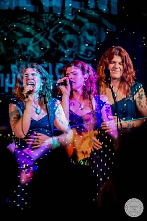 Three female singers performing together on stage at live music venue under colorful stage lighting.