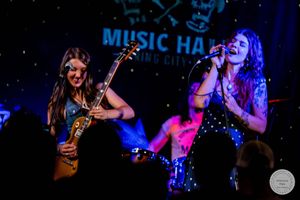 Two female musicians performing on stage at Music Hall, one playing guitar and one singing into a microphone.