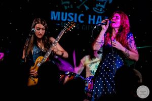 Two women rock performers on stage, guitarist in jeans and singer in polka dot dress under colorful lights.