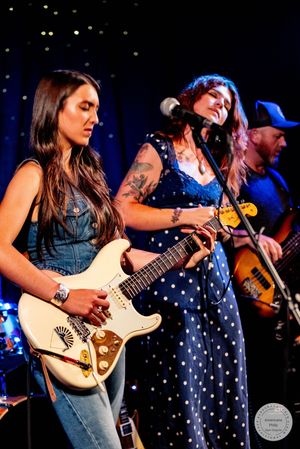 Two women playing electric guitars on stage, one in denim and one in blue polka dot dress, with male guitarist behind.