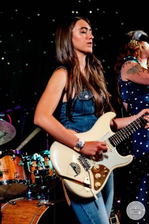 Young woman in denim overalls plays white electric guitar on stage with drummer visible behind her.