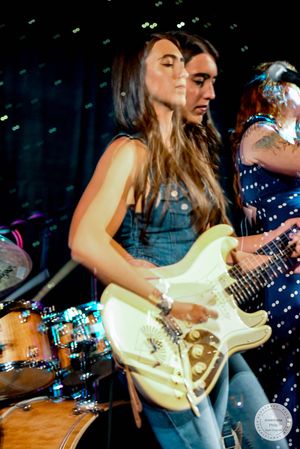 Female guitarist performing live on stage with white Stratocaster guitar beside another musician in blue polka dot dress.