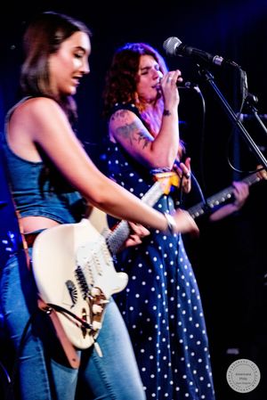 Two female musicians performing on stage, one playing white electric guitar in denim, other singing in polka dot dress.