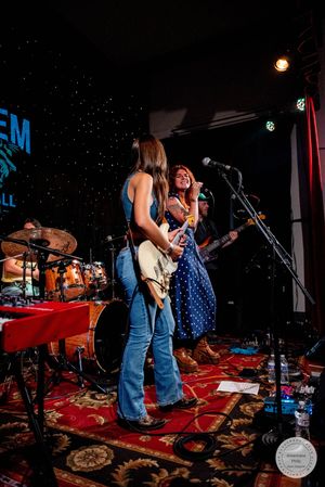 Two women perform on stage, one playing electric guitar in jeans, the other singing in a blue polka dot dress.