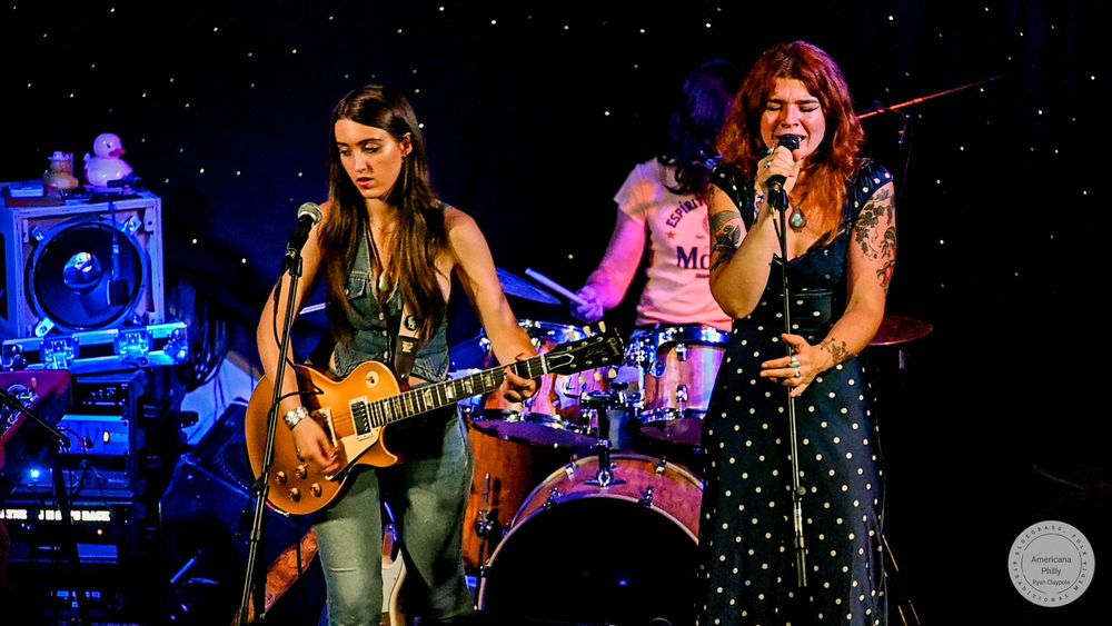 Two women performing on stage, one playing electric guitar and one singing into a microphone at a live music concert.
