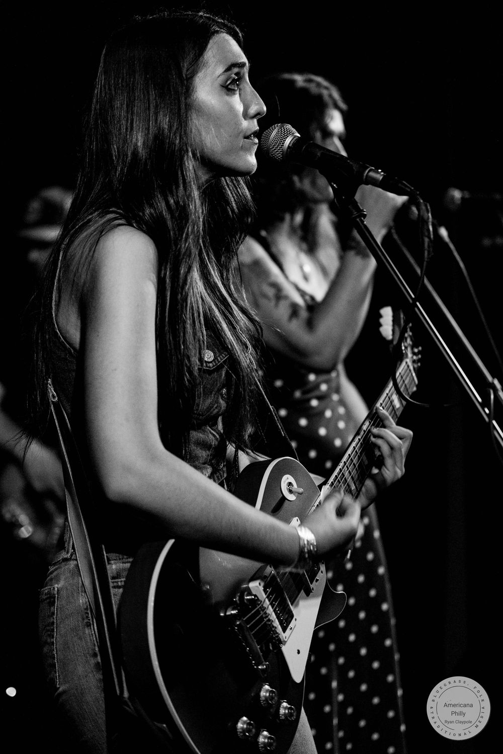 Woman playing electric guitar and singing into microphone on stage in black and white concert photo.