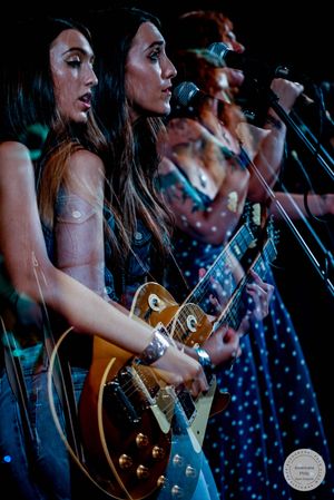 Four women perform on stage, with two playing electric guitars and singing into microphones at a live music concert.