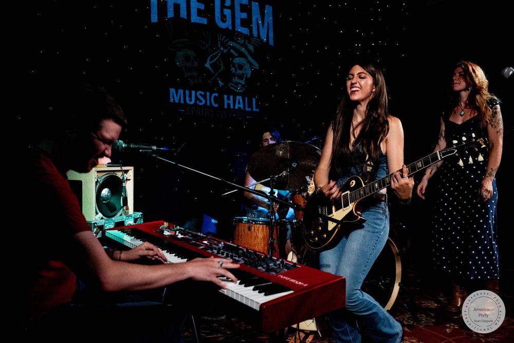 Band performing live at The Gem Music Hall, featuring a female guitarist, keyboardist, and bassist on a dimly lit stage.