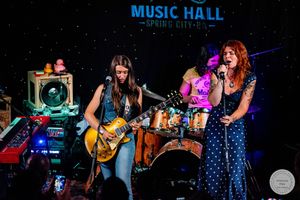 Two women performing live on stage at Music Hall Spring City, one playing Les Paul guitar under warm lights.