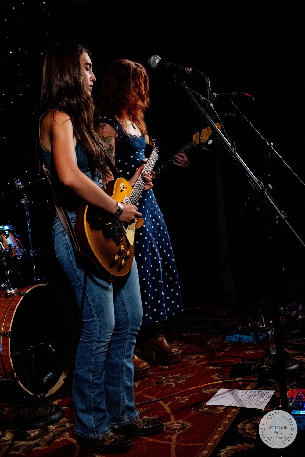 Two young women playing electric guitars on stage, one in jeans and one in a blue polka dot dress, performing at a live music event.